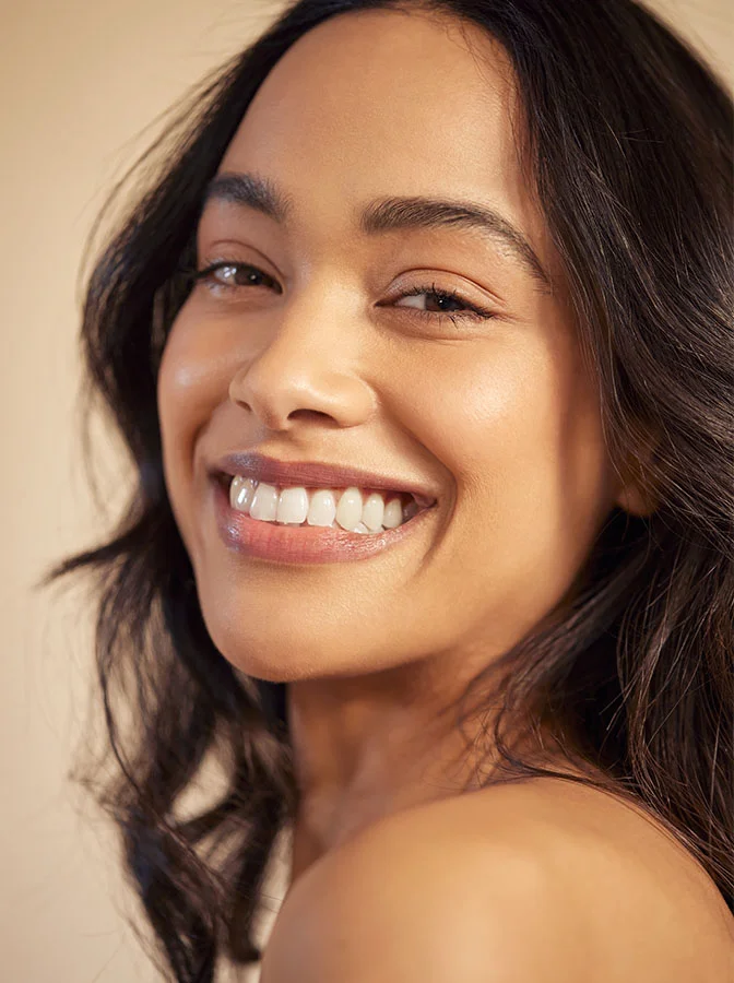 A smiling woman with radiant skin, showing her teeth, looking over her shoulder against a warm, neutral background - Brow Lift in Bala Cynwyd, PA