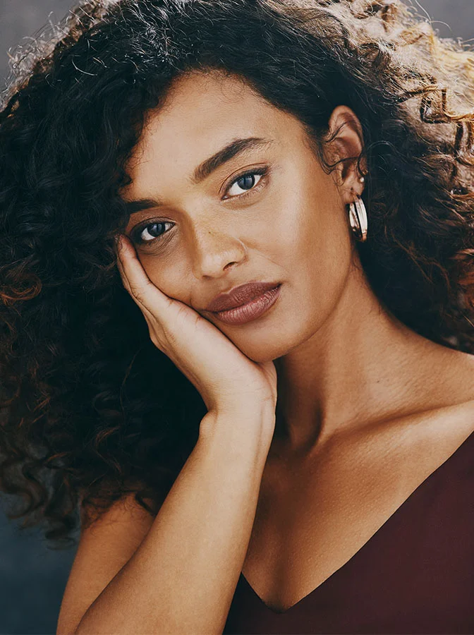 A woman with curly hair, resting her face on her hand, looking directly at the camera with a calm and reflective expression - Buccal Fat Removal in Bala Cynwyd, PA