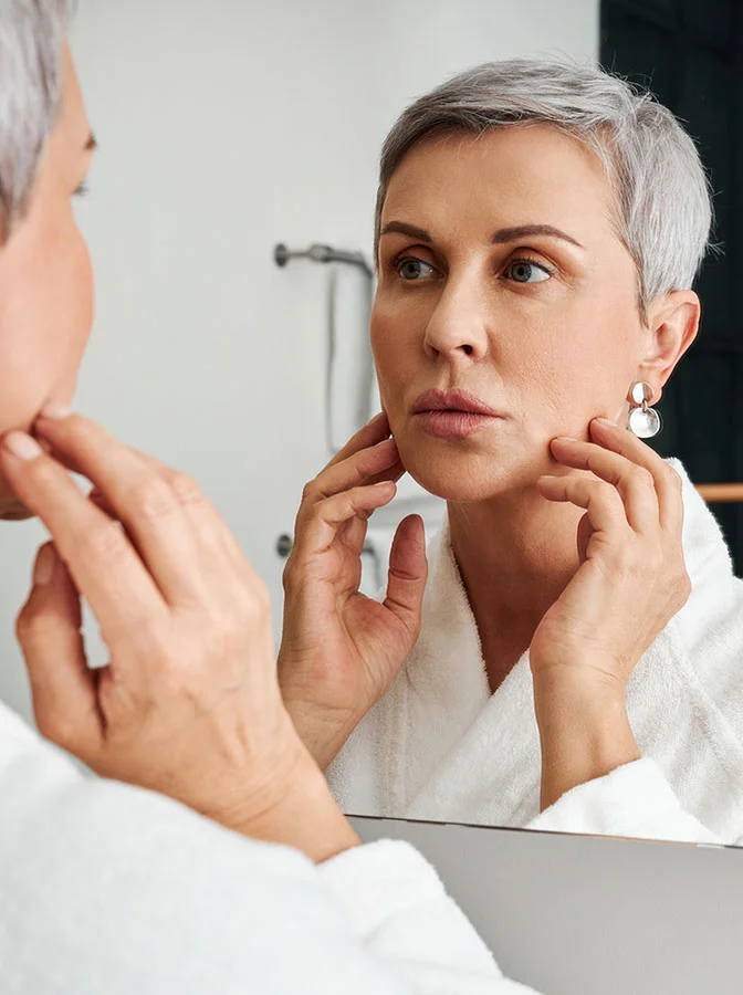 A woman with short gray hair, wearing a white bathrobe, examining her reflection in a mirror, gently touching her face - Chemical Peels in Bala Cynwyd, PA