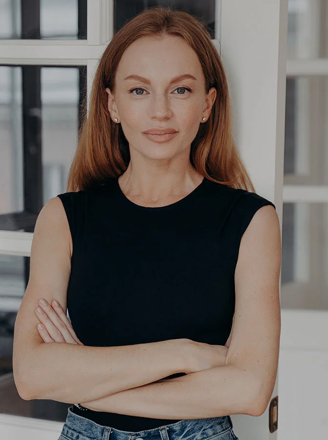 A woman standing confidently with her arms crossed, wearing a sleeveless black top, posing in front of a classic interior background with a soft smile- Direct Excision of Nasial Labial Folds in Bala Cynwyd, PA