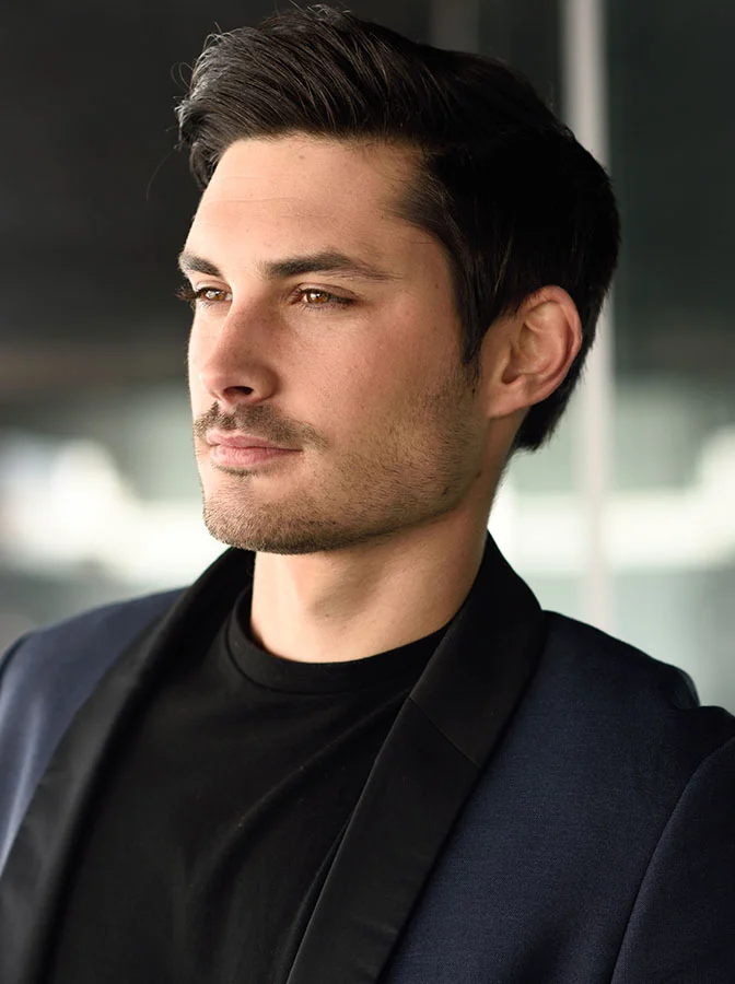 A man with short hair and light facial hair, wearing a dark suit and black shirt, looking confidently into the distance in a professional setting - Earlobe Repair Reduction in Bala Cynwyd, PA