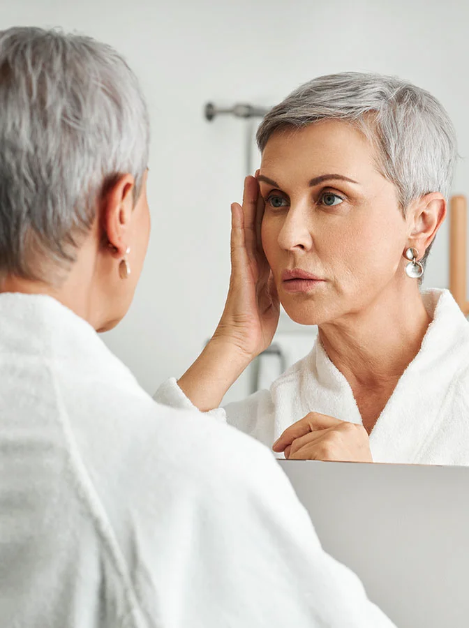 Mature woman with short silver hair looking in the mirror, gently touching her face, reflecting on her appearance with a calm expression - Facial Fat Transfer in Bala Cynwyd, PA