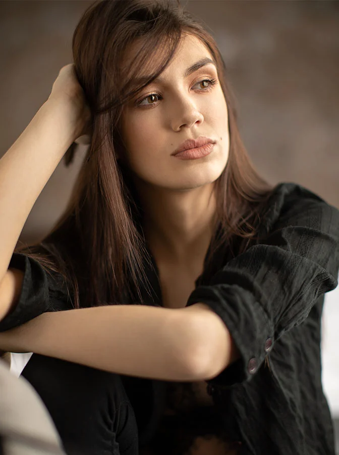 A woman with long dark hair sitting pensively, resting her hand on her head, wearing a black top in soft, natural lighting - Lip Augmentation in Bala Cynwyd, PA