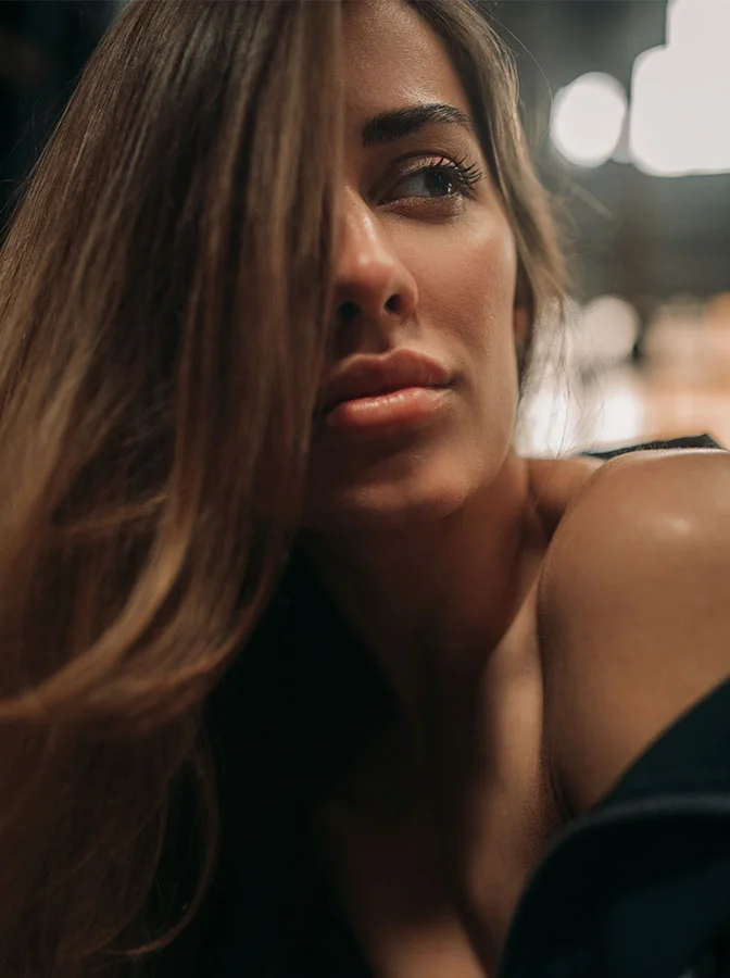 A close-up shot of a woman with long brunette hair, gazing off to the side, her hair partially covering her face, in a softly lit environment - Lip Lift in Bala Cynwyd, PA