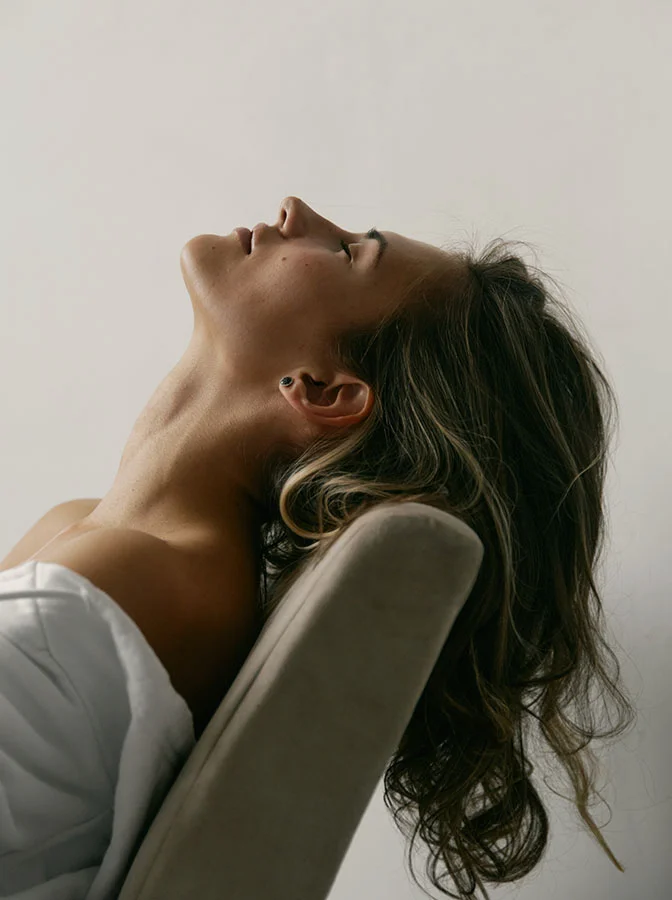 Woman reclining in a beige chair, head tilted back with eyes closed, basking in soft natural light - Necklift in Bala Cynwyd, PA