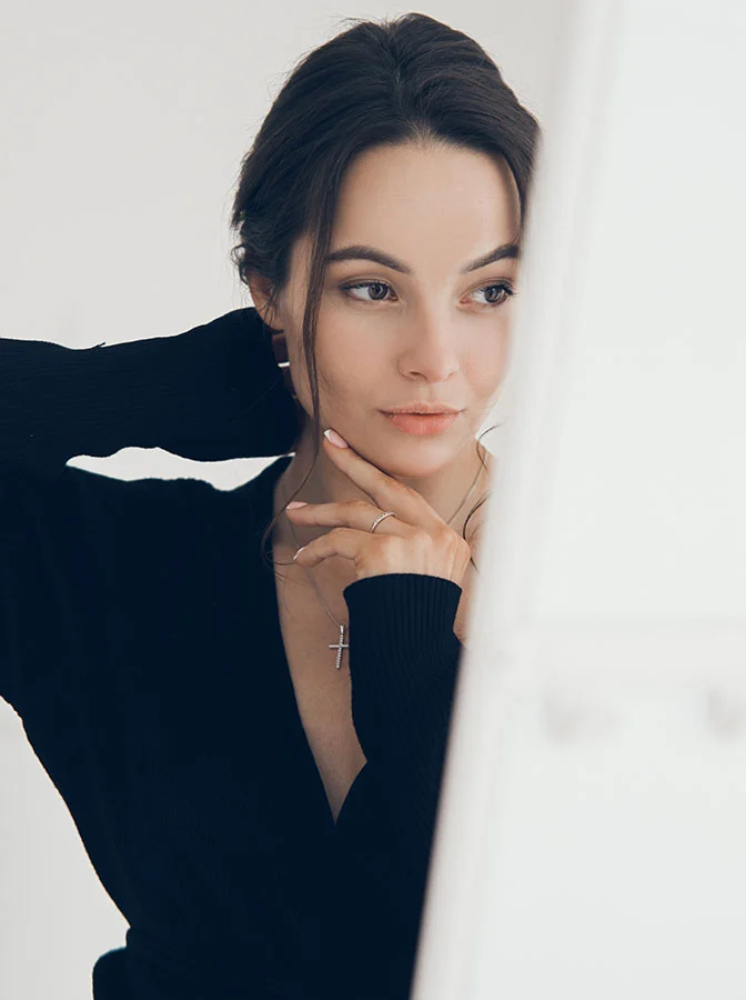 A woman with her hand resting near her chin, wearing a black top, looking thoughtfully into a mirror, with soft lighting on her face - Skin Resurfacing Pixel Laser in Bala Cynwyd, PA